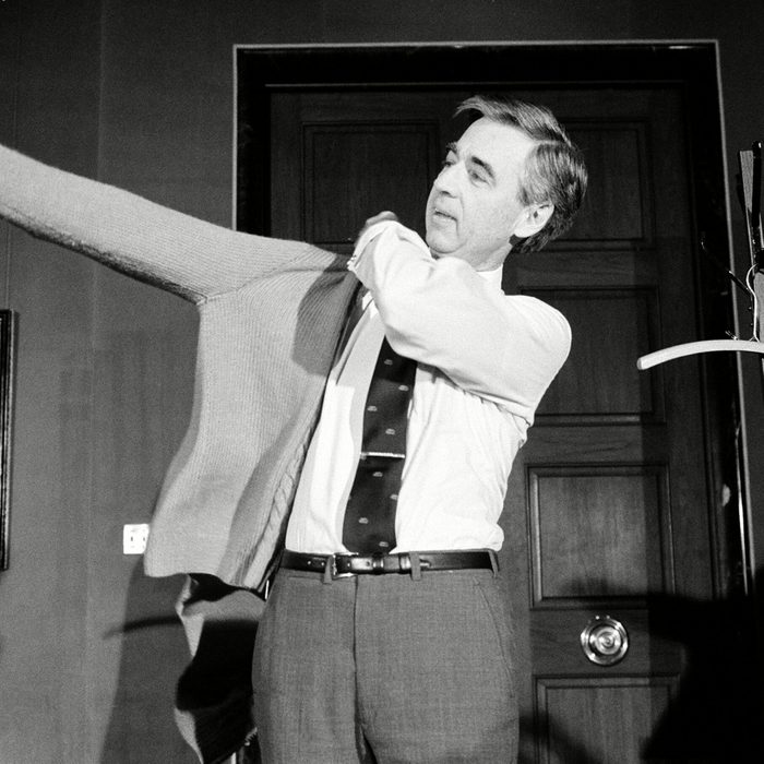 Man puts on sweater in an office with coat rack and painting; another man observes.