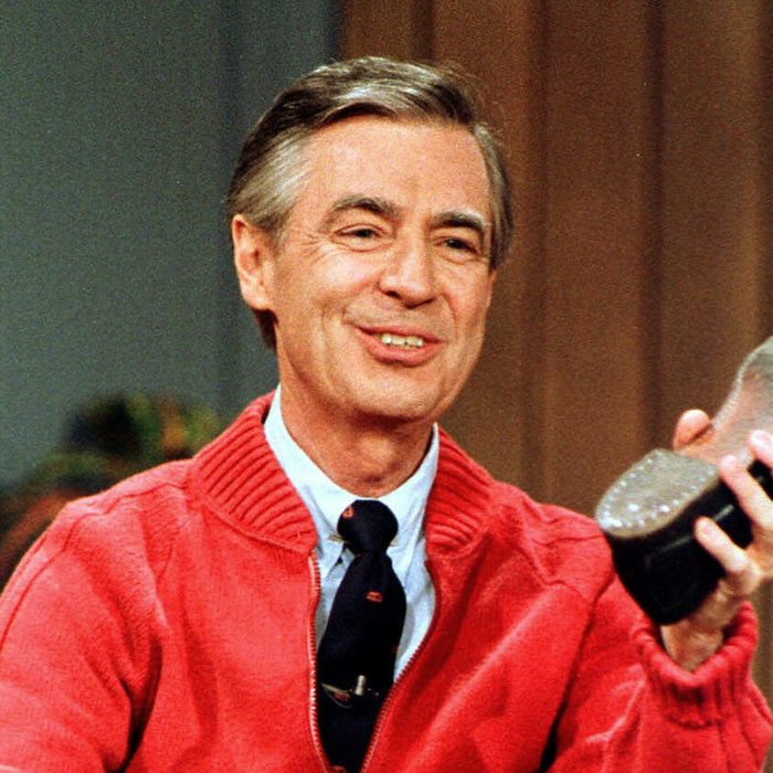 Man holding shoe, smiling; wearing a red sweater and tie, seated indoors with curtains and plants in the background.