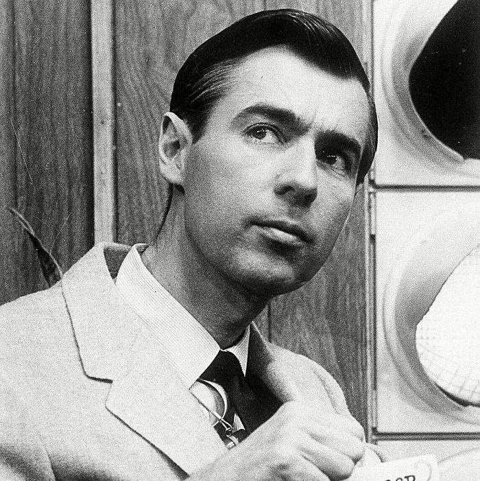 Man holding a sign with text, "WON'T YOU BE MY NEIGHBOR MISTER ROGERS," indoors, against a wooden background with a circular object nearby.