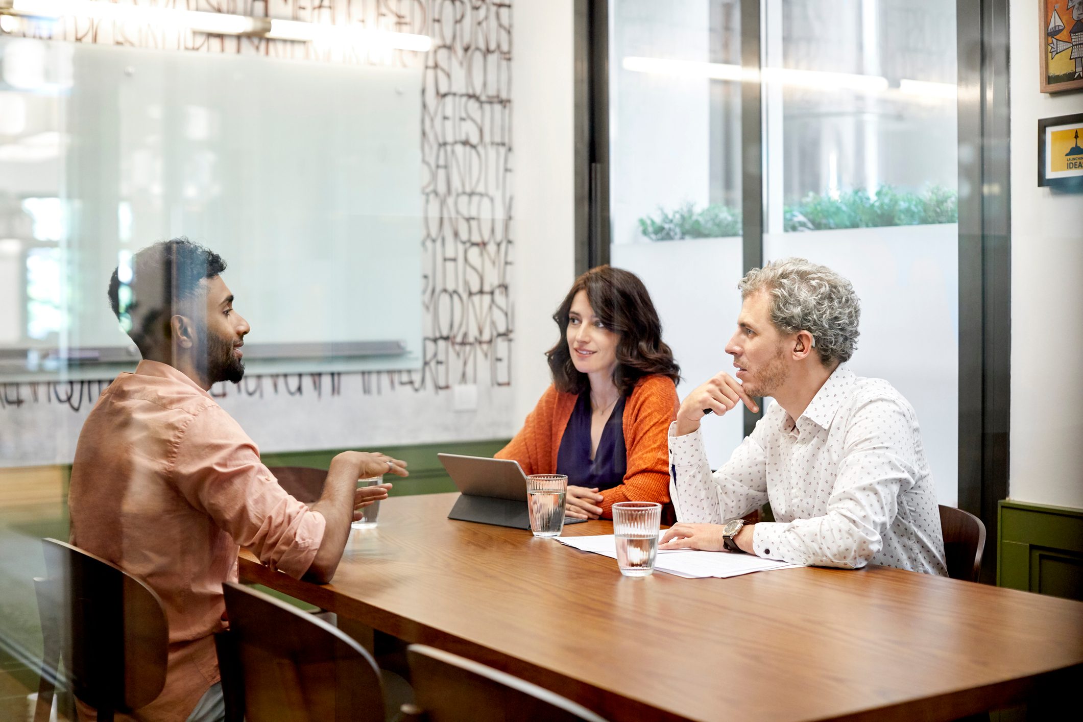 Three people discuss around a wooden table in a modern office, with a laptop and papers. A glass wall displays abstract lettering.