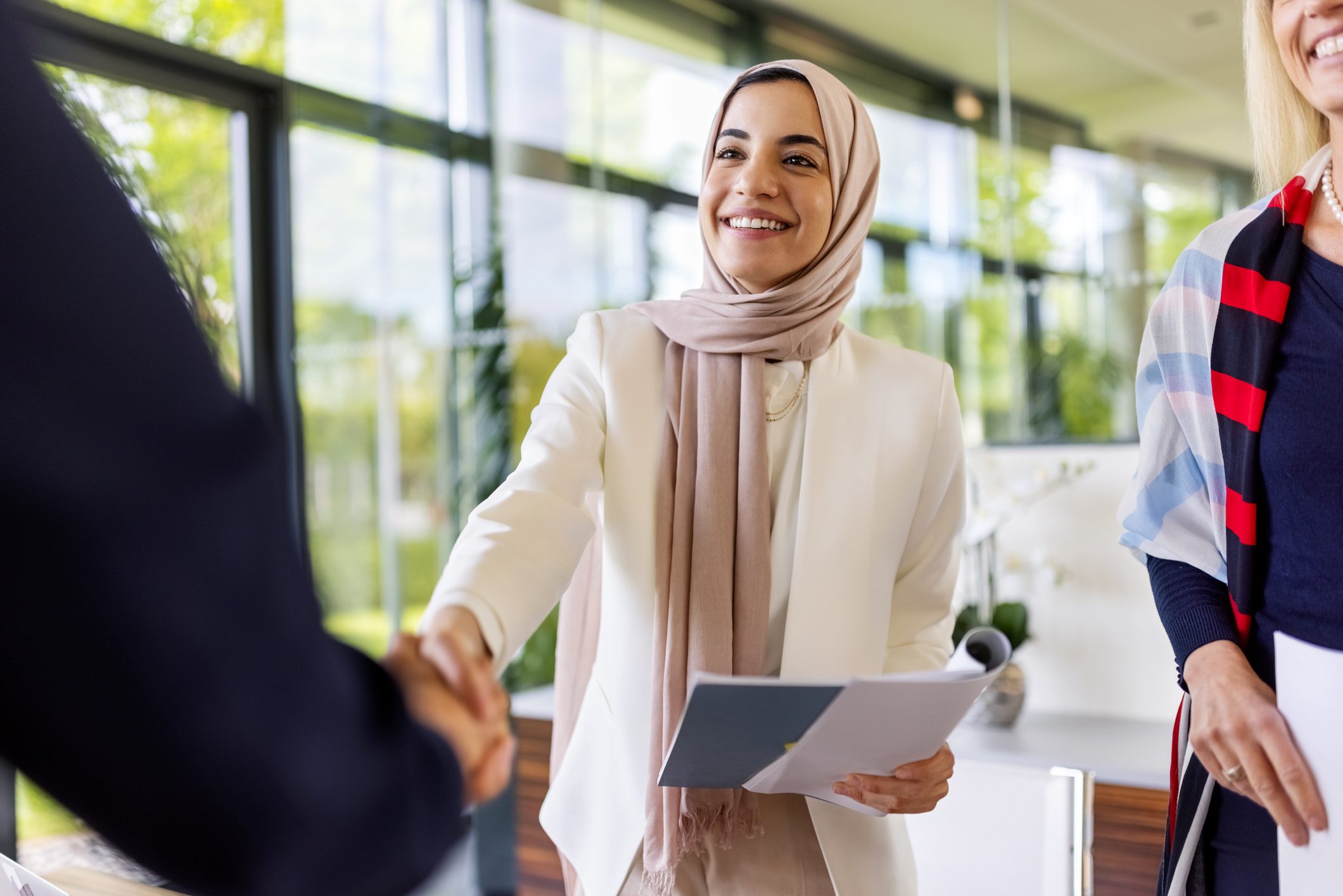 A woman in a hijab smiling, shaking hands, holds documents in a bright, modern office setting.