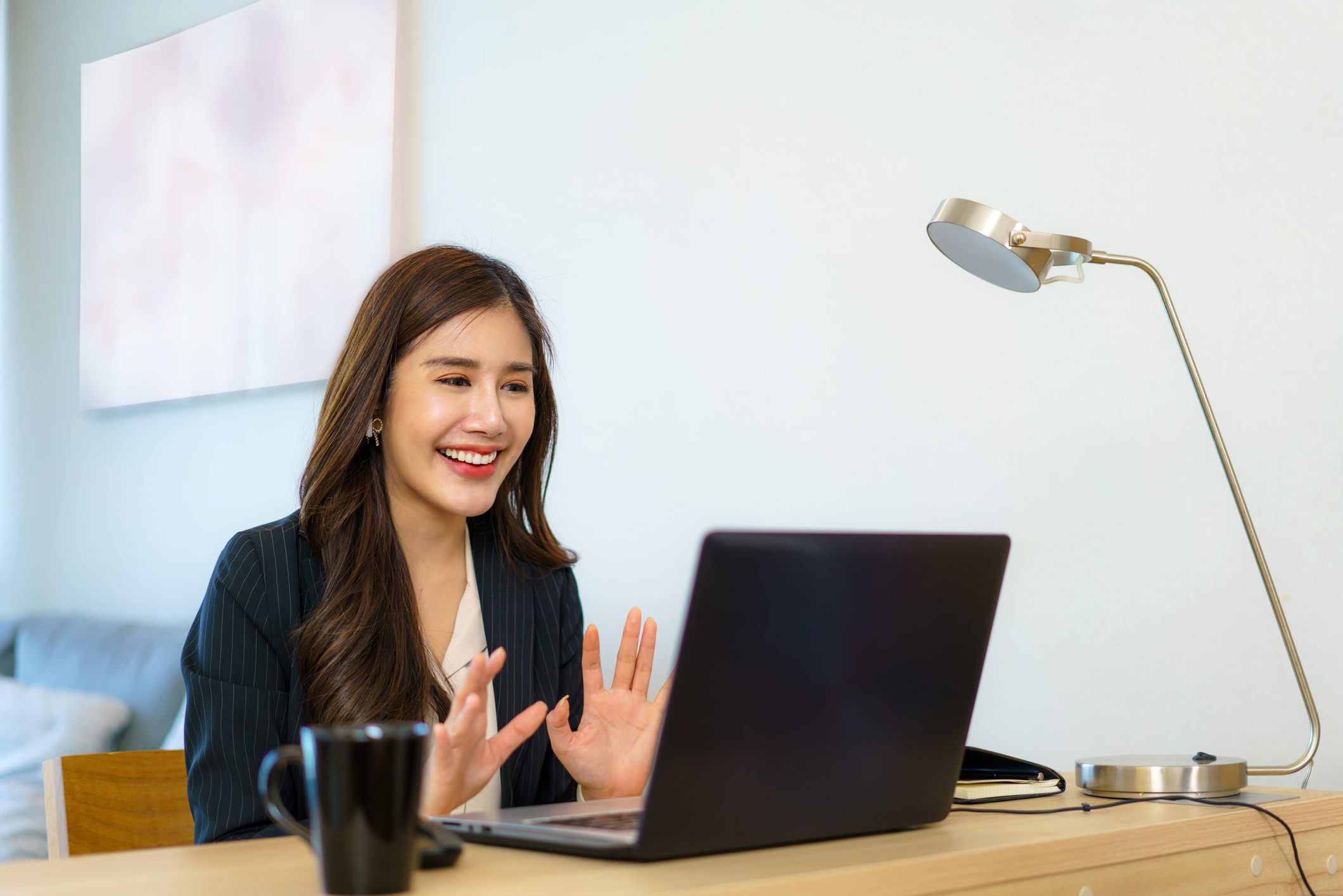 A woman smiles and gestures during a video call, seated at a desk with a laptop, mug, and lamp in a bright room.