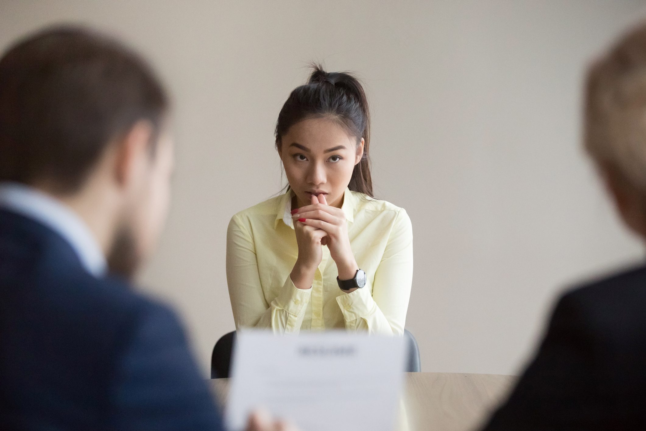 Woman sits thoughtfully at a table, hands together, during a job interview with two blurred-in-the-foreground interviewers.