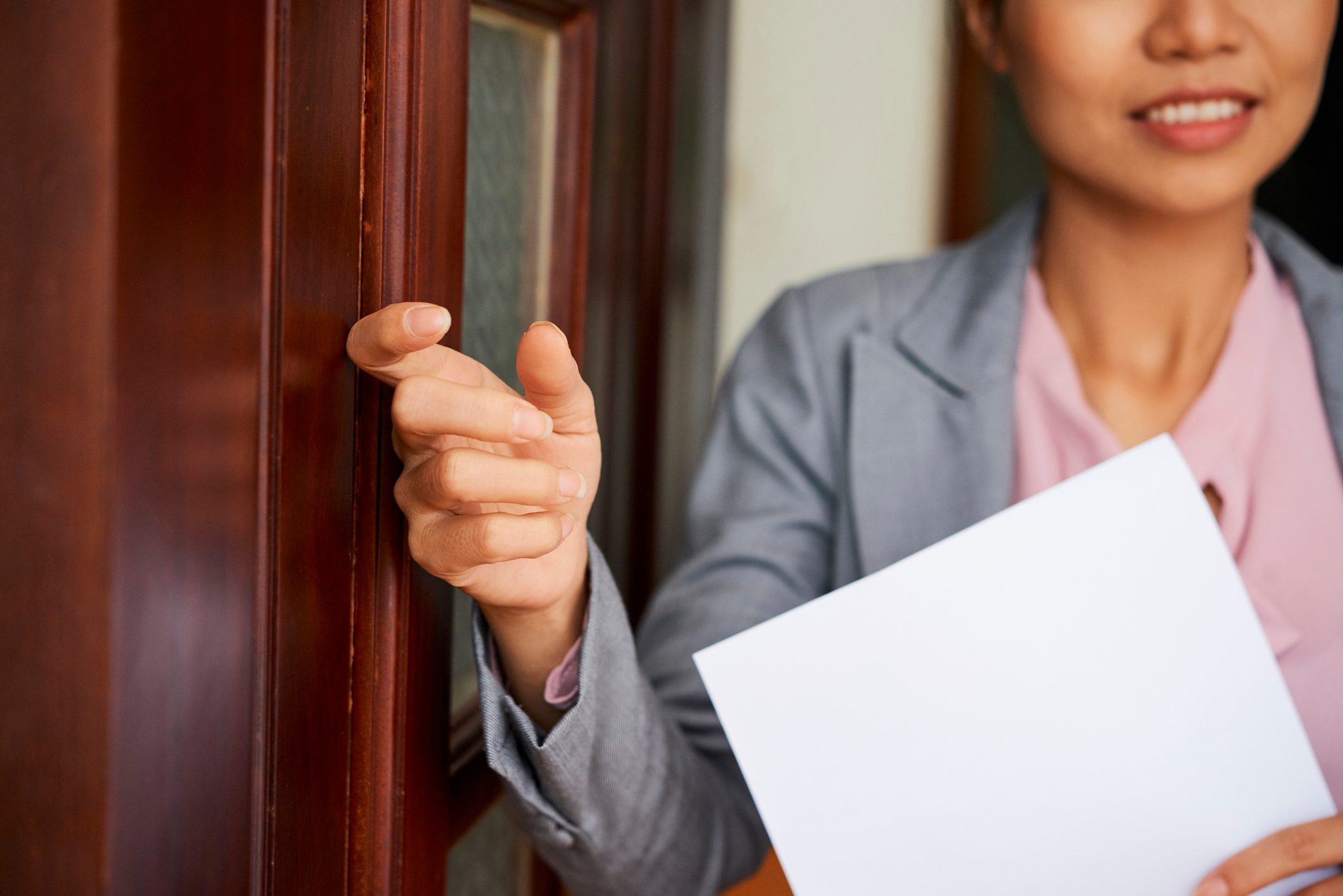 Hand knuckles on a wooden door, person holding a blank white paper, wearing a gray blazer, slightly smiling, indoors.