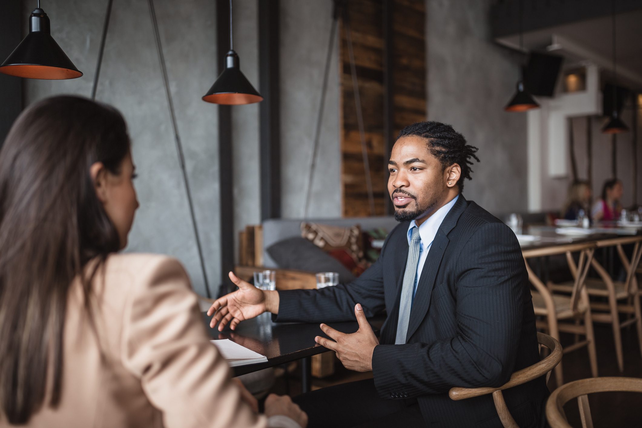Man talking across table; both in business attire, discussing in a modern cafÃ© with industrial lighting and wooden decor.