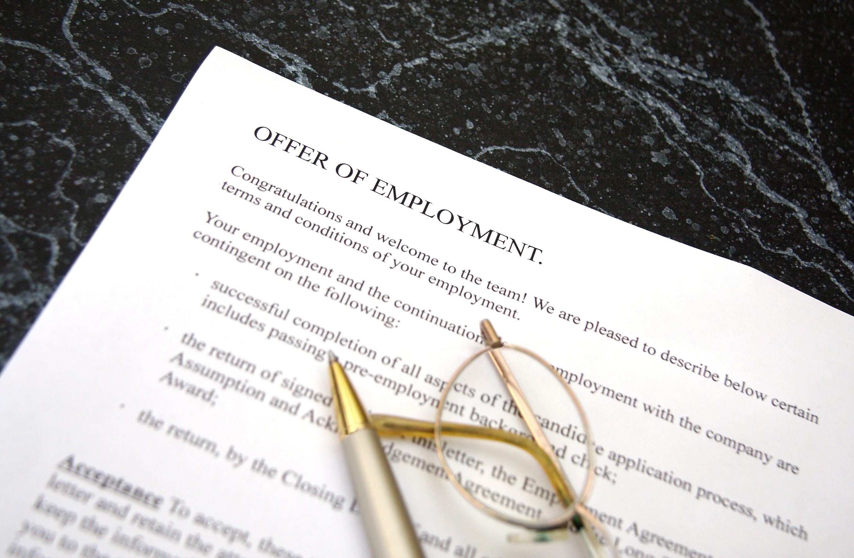 Employment offer letter lies on dark marble surface, accompanied by a pen and eyeglasses, within professional and formal setting.