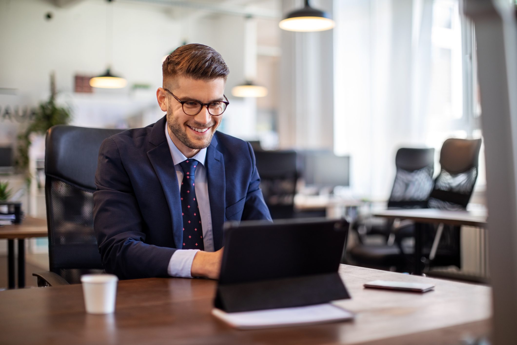 A man in a suit smiles while working on a tablet in a modern office with large windows and natural light.