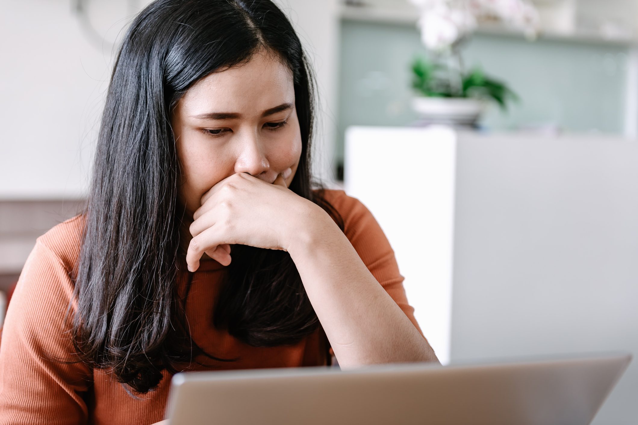 A person thinks deeply, hand on face, while looking at a laptop in a softly lit room with a plant nearby.