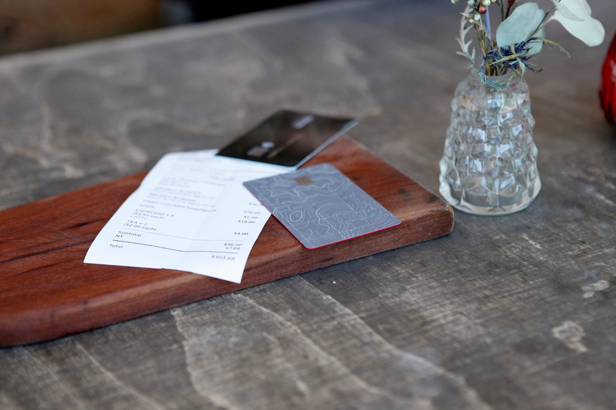 Receipt, credit card, and paper on wooden board; small vase with flowers sits nearby on a textured table.