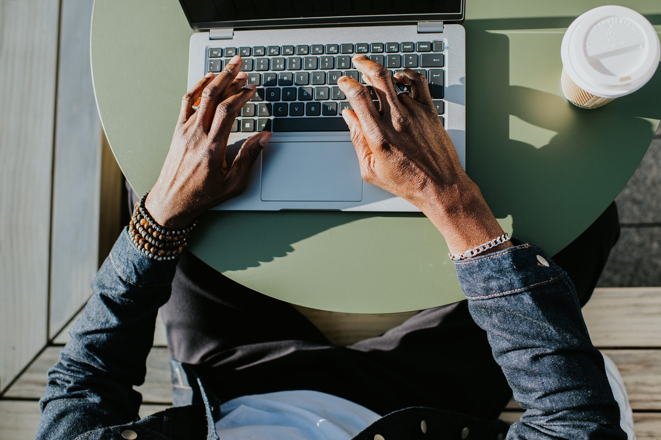 Hands type on a laptop, positioned on a green table outdoors, with a coffee cup nearby.