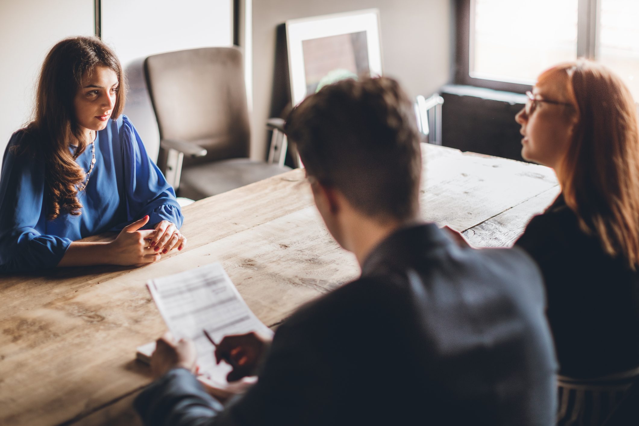 Woman sits attentively, engaged in conversation with two people, across a wooden table in a bright office setting.