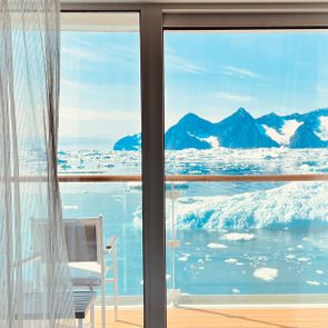 Chair sits on a balcony overlooking a glacial ocean scene with snow-capped mountains in the distance, framed by a glass door with curtain.