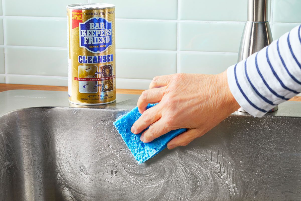 Hand cleaning stainless steel sink with a blue sponge in a circular motion; bar keeper's friend container nearby on the edge of the sink