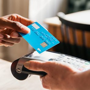 A hand holds a credit card above a payment terminal at a cafe table with a cappuccino cup.