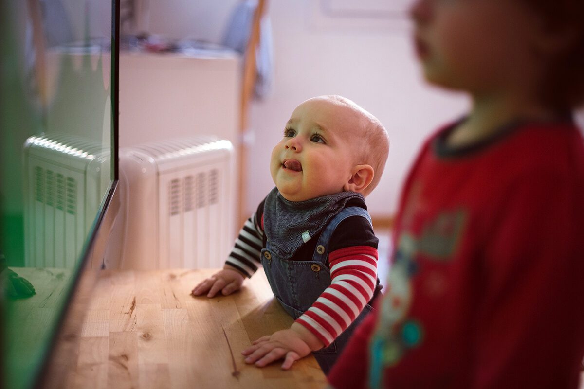 A baby in striped sleeves stands at a wooden surface, looking up and smiling at something off-camera. Another child in a red shirt is blurred in the foreground.